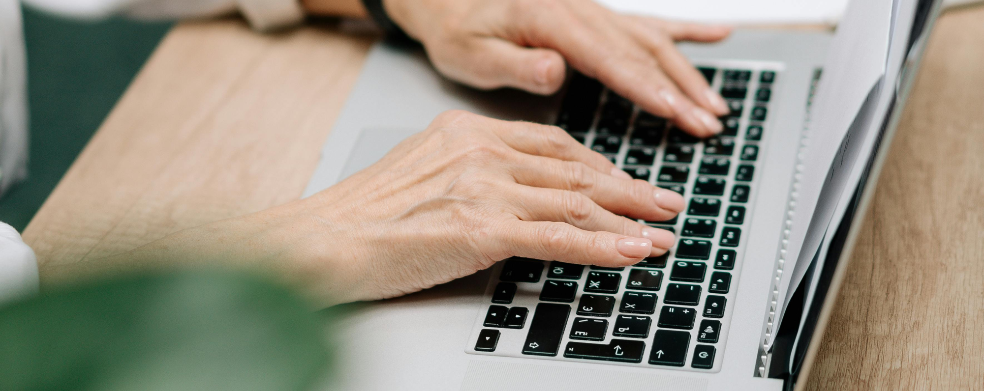 An adult typing on a laptop at a desk, in a well-lit office space with papers and plants nearby.